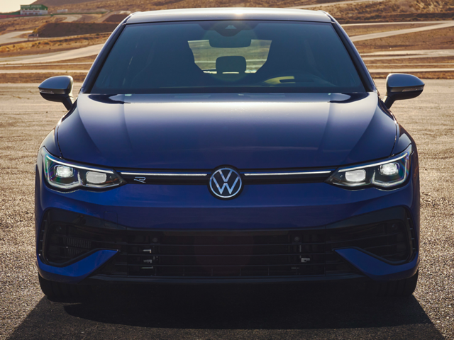 A straight-on view of a blue 2024 Volkswagen Golf-R sitting in a lot backed by winding wilderness roads. 