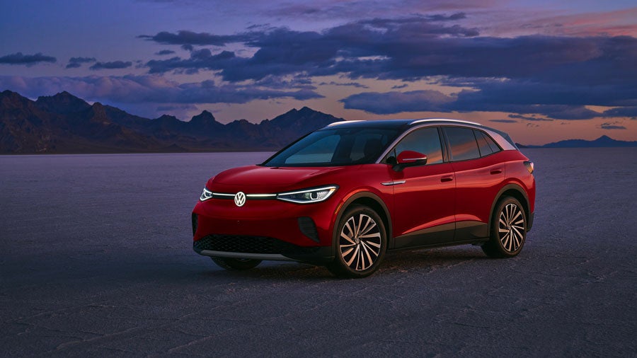 A ¾ front of an ID.4 SUV in Aurora Red Metallic in a natural sunset setting. The illuminated front Volkswagen logo and connecting line glows.